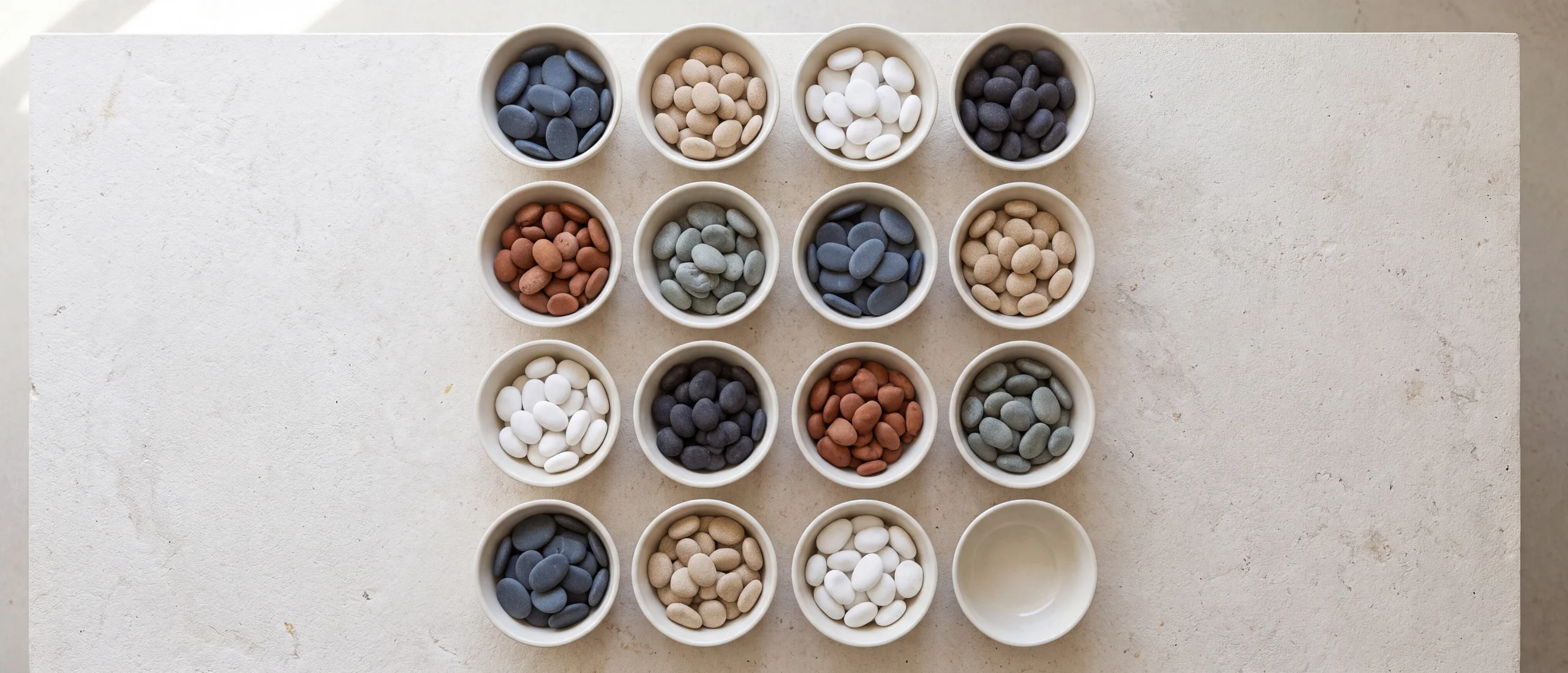 Sixteen shallow off-white dishes in a four by four grid on a pale limestone table, fifteen holding smooth pebbles sorted by tone and one at the lower right still empty.